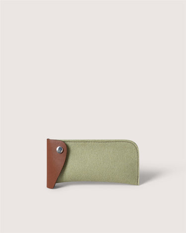 A slim, minimalist card holder or wallet made of sage green felt with a contrasting brown leather tab on the left side, secured by a silver snap button, set against a solid light beige background.