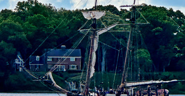 Schooner Sultana sailing down a river with a house in the background.