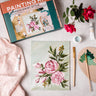 Floral painting kit with completed artwork, paintbrushes, and supplies on a light surface.
