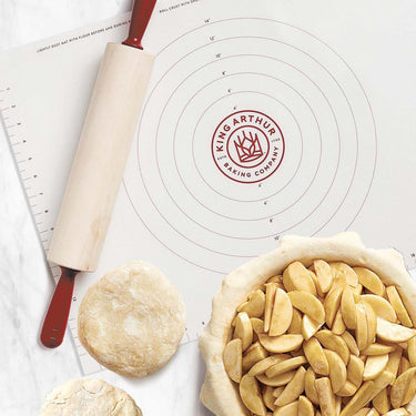 Baking setup with a rolling pin, pie crust, and apples on a King Arthur Baking Company mat.