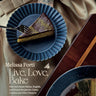 Cover features a woman in a black top, and a taupe colored satin skirt holding a blue ruffled edge plate with a chocolate iced piece of cake. She is holding a fork in her right hand. On the dark wood table in front of her is a platter with the rest of the cake with her slice taken out.